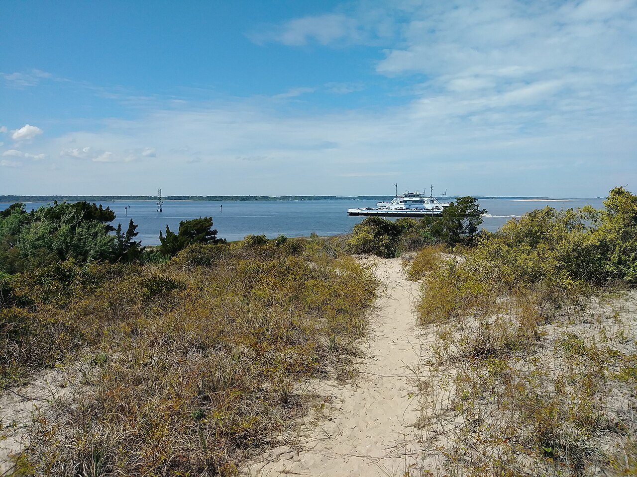 A sandy path through the dunes to the water, with a ferry passing offshore.